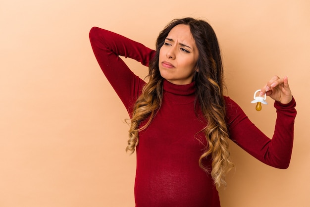 Giovane donna incinta messicana che tiene il ciuccio isolato sulla parete beige che tocca la parte posteriore della testa, pensando e facendo una scelta.