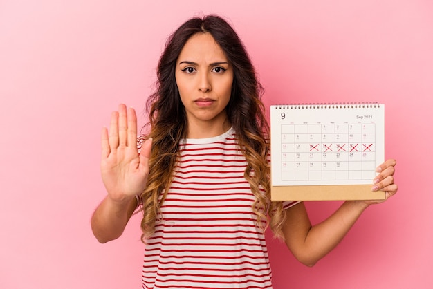 Young mexican woman holding a calendar isolated on pink wall standing with outstretched hand showing stop sign, preventing you.