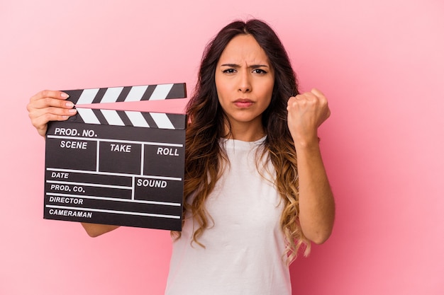 Young mexican woman holding clapperboard isolated on pink background showing fist to camera, aggressive facial expression.