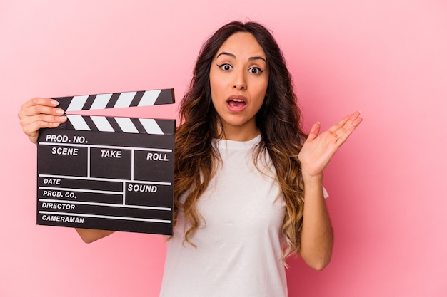 Young mexican woman holding clapperboard isolated on pink background surprised and shocked.