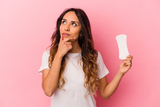 Giovane donna messicana che tiene un impacco isolato su sfondo rosa guardando lateralmente con espressione dubbiosa e scettica.