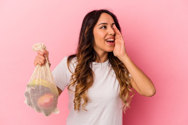 Молодая мексиканская женщина держит сумку с фруктами, изолированную на розовом фоне, кричит и держит ладонь возле открытого рта.