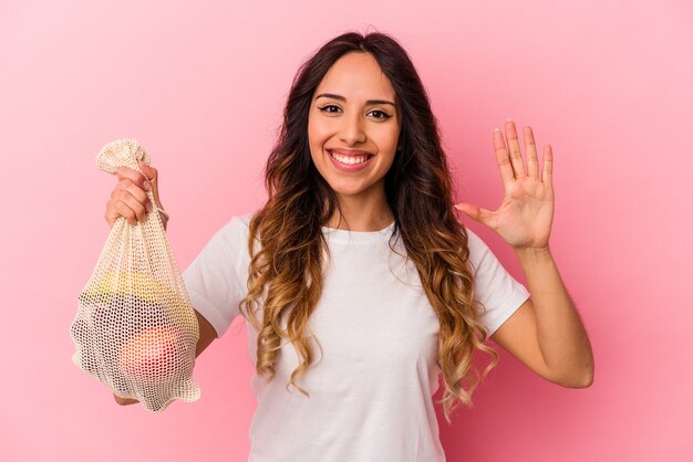 Young mexican woman holding a fruit bag isolated on pink background smiling cheerful showing number five with fingers.