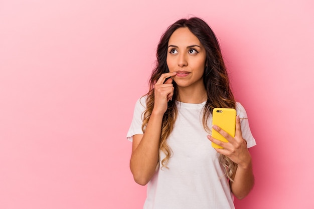 Young mexican woman holding a mobile phone isolated on pink background relaxed thinking about something looking at a copy space.