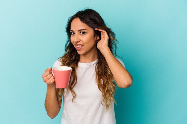 Young mexican woman holding a mug isolated on blue background trying to listening a gossip.