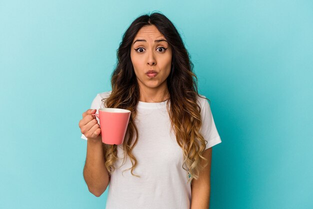 Young mexican woman holding a mug isolated on blue wall shrugs shoulders and open eyes confused.