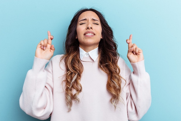 Young mexican woman isolated on blue background crossing fingers for having luck