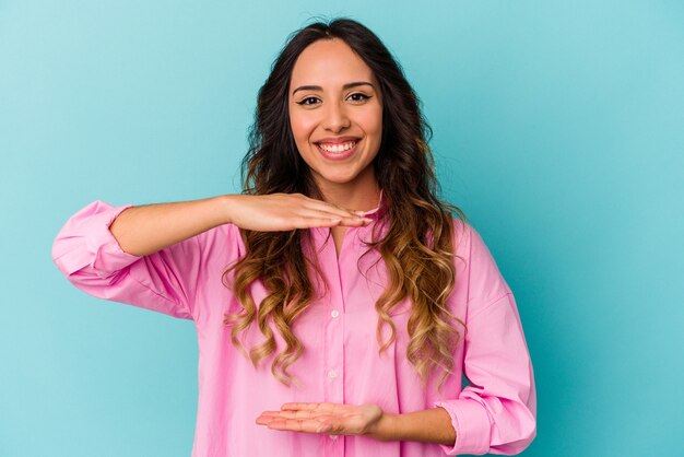 Young mexican woman isolated on blue background holding something with both hands, product presentation.