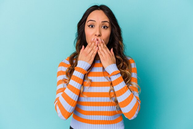 Young mexican woman isolated on blue background shocked covering mouth with hands.