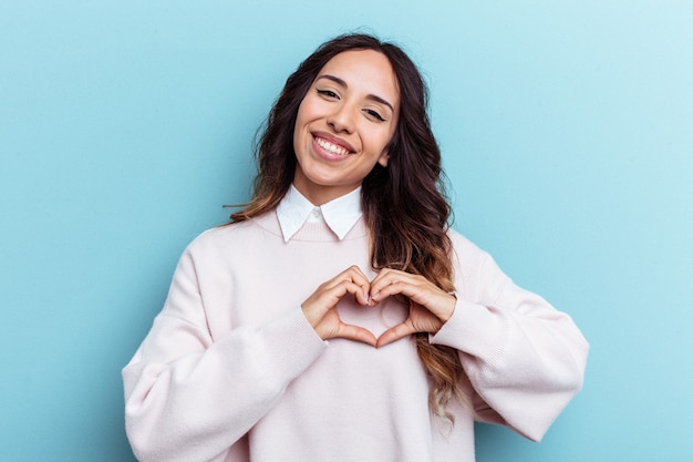 Young mexican woman isolated on blue background smiling and showing a heart shape with hands
