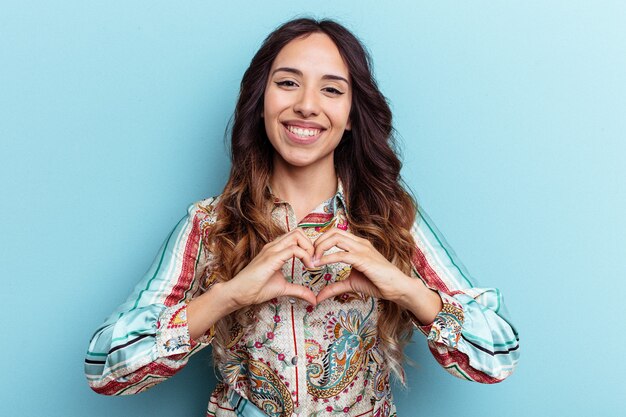 Young mexican woman isolated on blue background smiling and showing a heart shape with hands.