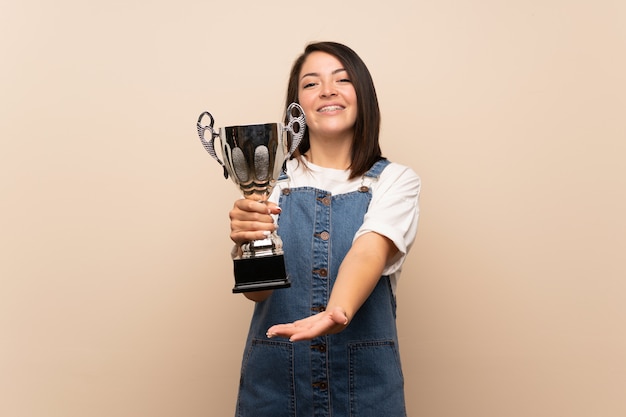 Young Mexican woman over isolated wall holding a trophy