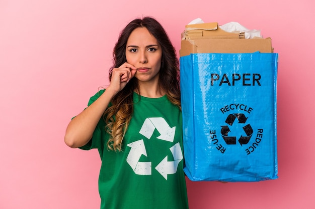 Young mexican woman recycling cardboard isolated on pink background with fingers on lips keeping a secret.