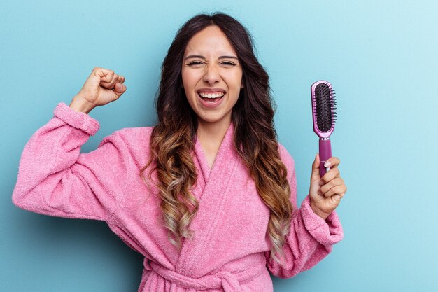 Young mexican woman wearing a bathrobe holding a brush isolated on blue background raising fist after a victory, winner concept.