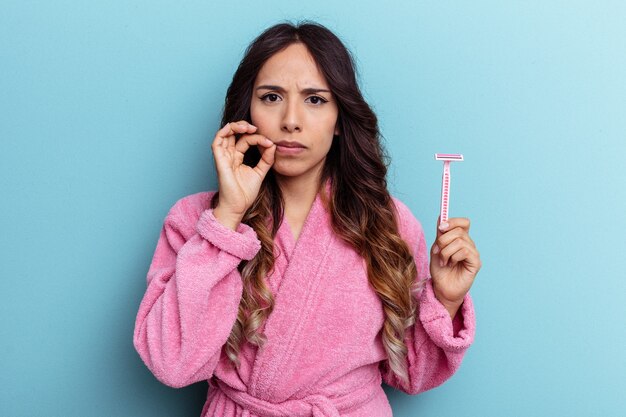 Young mexican woman wearing a bathrobe holding a knife isolated on blue background with fingers on lips keeping a secret.