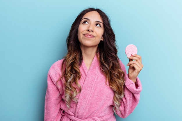 Young mexican woman wearing a bathrobe holding a make-up
remover sponge isolated on blue background dreaming of achieving
goals and purposes