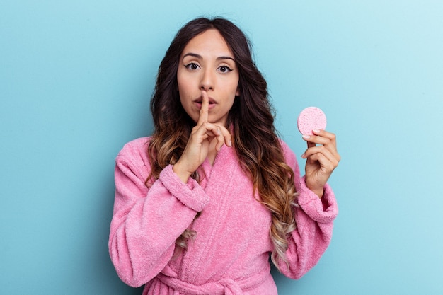 Young mexican woman wearing a bathrobe holding a make-up\
remover sponge isolated on blue background keeping a secret or\
asking for silence.