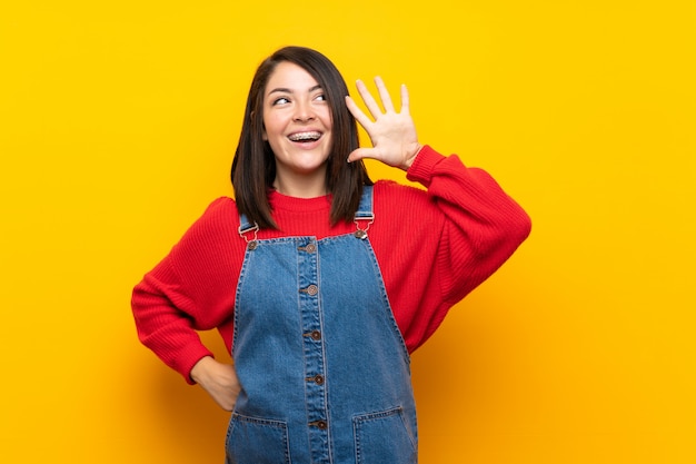 Young mexican woman with overalls over yellow wall listening to something by putting hand on the ear