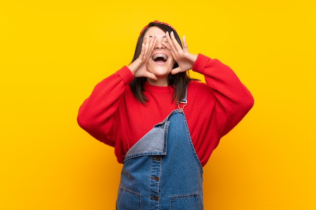 Young Mexican woman with overalls over yellow wall shouting and announcing something