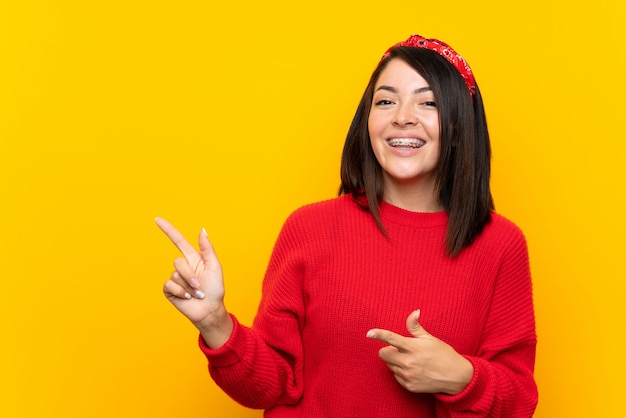 Young Mexican woman with red sweater over yellow wall pointing finger to the side