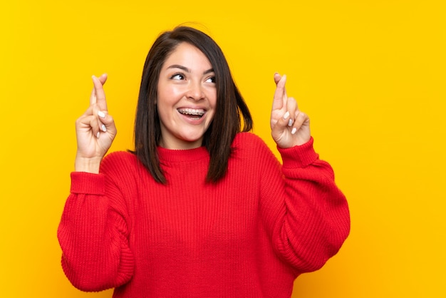 Young mexican woman with red sweater over yellow wall with fingers crossing