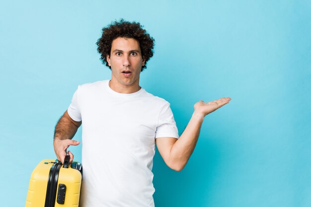 Young middle aged man holding a suitcase impressed holding copy space on palm.