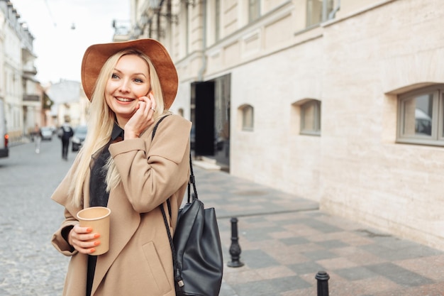 사진 가 코트와 모자를 입고 젊은 밀레니엄 웃는 금발 여자. 유럽 거리에서 전화 통화를 하고 커피 한 잔을 들고 있는 귀여운 소녀.
