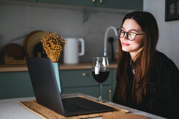 Giovane donna millenaria che ha videochiamata sul computer portatile e beve vino, usa la tecnologia per comunicare con amici o familiari