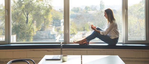 A young millennial woman looks in a smartphone while sitting by the window in a bright interior. Banner.