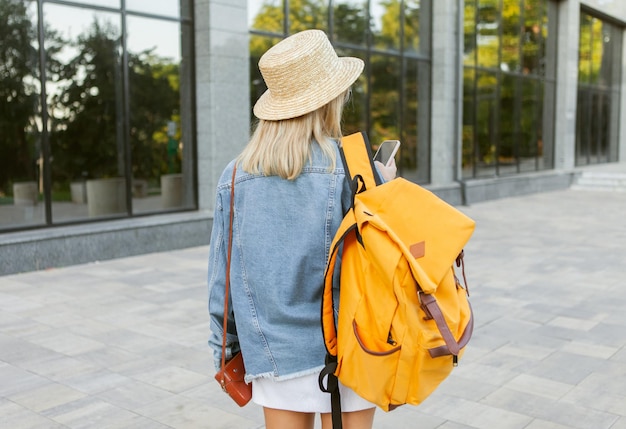 Photo young millennial woman tourist in hat with tourist backpack uses smartphone while sitting on a bench at city park. back view