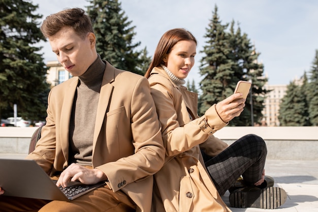 Young millennials in elegant beige trenchcoats, pullovers and pants using mobile gadgets outdoors while sitting against evergreen trees