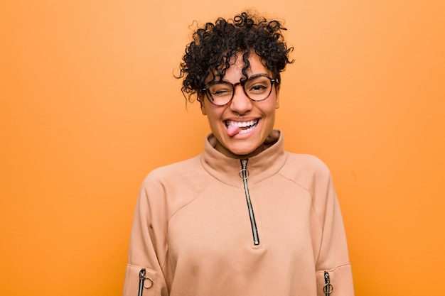 Young mixed african american woman against a brown wall funny and friendly sticking out tongue.