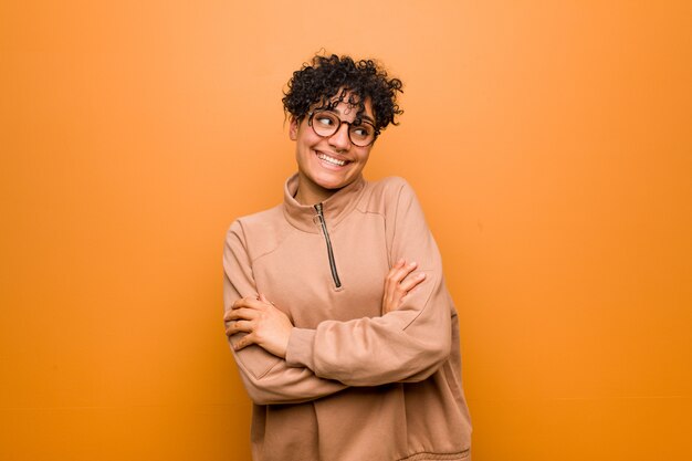 Young mixed african american woman against a brown wall smiling confident with crossed arms.