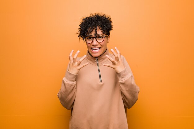 Young mixed african american woman against a brown wall upset screaming with tense hands.