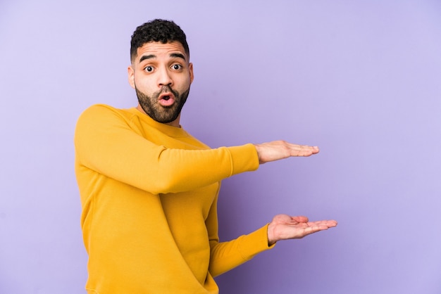 Young mixed race arabic man isolated shocked and amazed holding a copy space between hands.