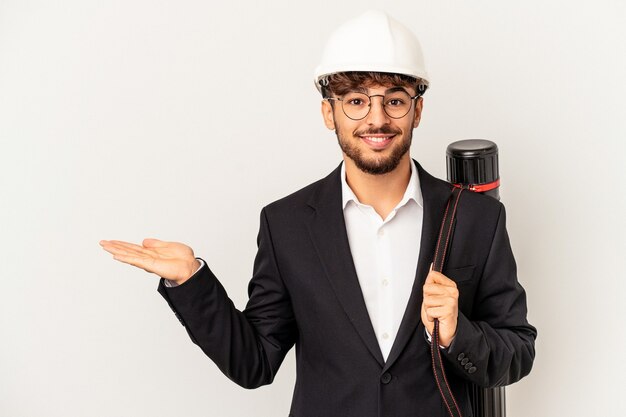 Young mixed race architect man wearing a helmet isolated on grey background showing a copy space on a palm and holding another hand on waist.