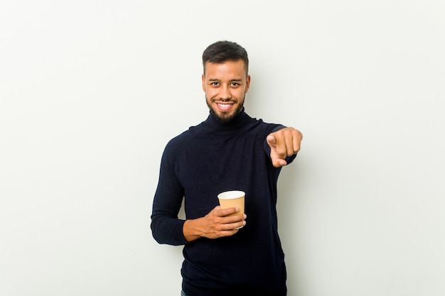 Young mixed race asian man holding a takeaway coffee cheerful smiles pointing to front.
