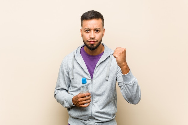 Young mixed race asian man holding a water bottle showing fist to camera, aggressive facial expression.