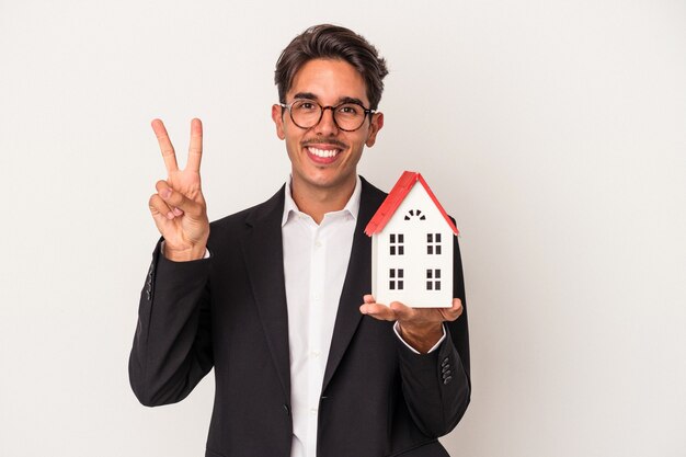 Young mixed race business man holding a toy house isolated on white background joyful and carefree showing a peace symbol with fingers.