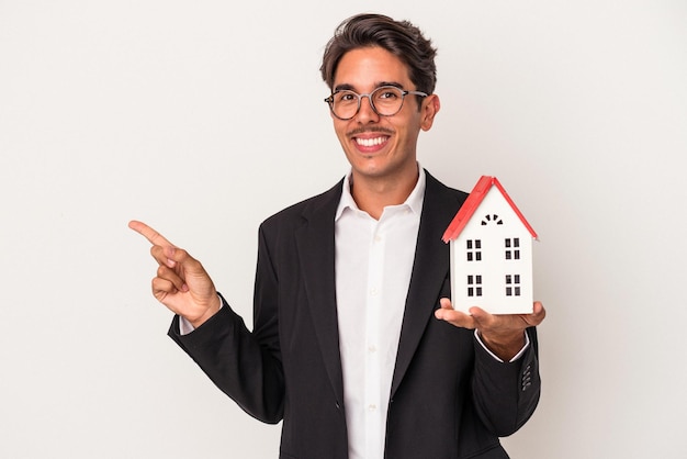 Young mixed race business man holding a toy house isolated on white background smiling and pointing aside, showing something at blank space.