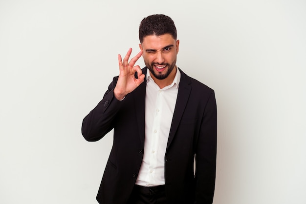 Young mixed race business man isolated on white winks an eye and holds an okay gesture with hand.