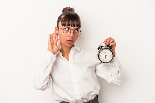 Young mixed race business woman holding an alarm clock isolated on white background  trying to listening a gossip.