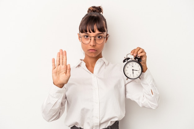写真 白い背景で隔離の目覚まし時計を保持している若い混血ビジネス女性は、一時停止の標識を示す伸ばした手で立って、あなたを防ぎます。