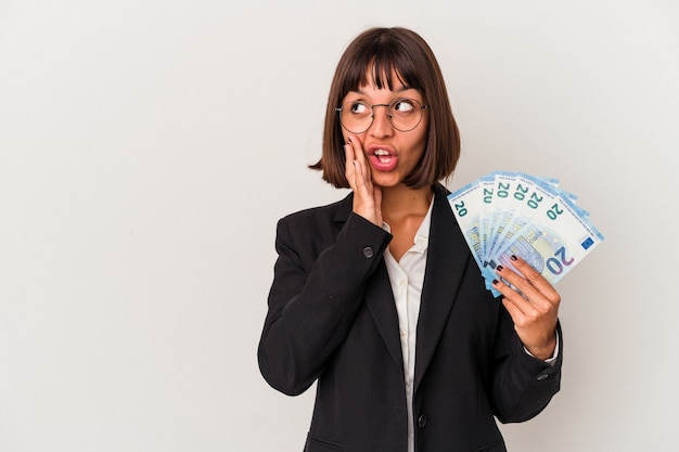 Young mixed race business woman holding a banknotes isolated on white background is saying a secret hot braking news and looking aside