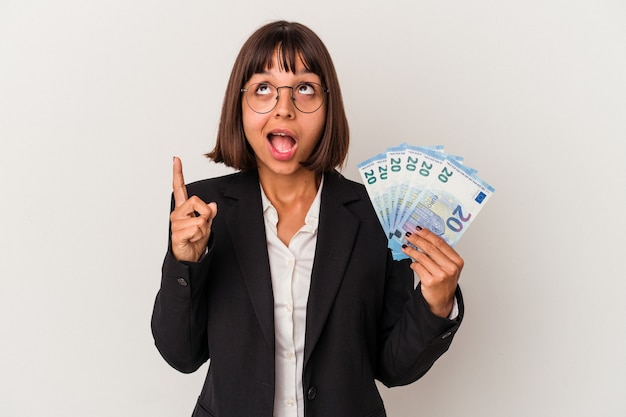 Young mixed race business woman holding a banknotes isolated on white background pointing upside with opened mouth.