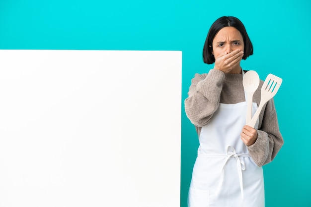 Young mixed race cook woman with a big placard isolated on blue background covering mouth with hands