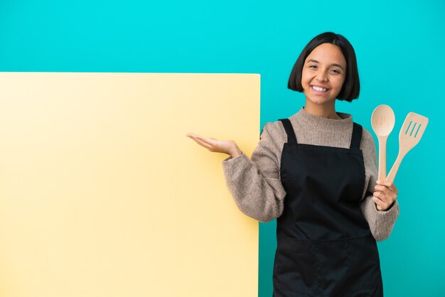 Giovane donna cuoca di razza mista con un grande cartellone isolato su sfondo blu che tiene immaginario copyspace sul palmo per inserire un annuncio