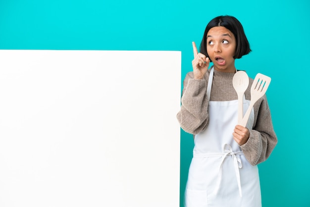 Young mixed race cook woman with a big placard isolated on blue background intending to realizes the solution while lifting a finger up