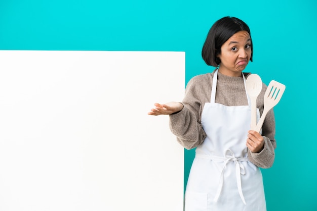 Young mixed race cook woman with a big placard isolated on blue background making doubts gesture while lifting the shoulders