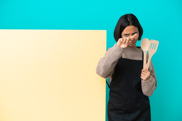 Young mixed race cook woman with a big placard isolated on blue background making stop gesture with her hand to stop an act